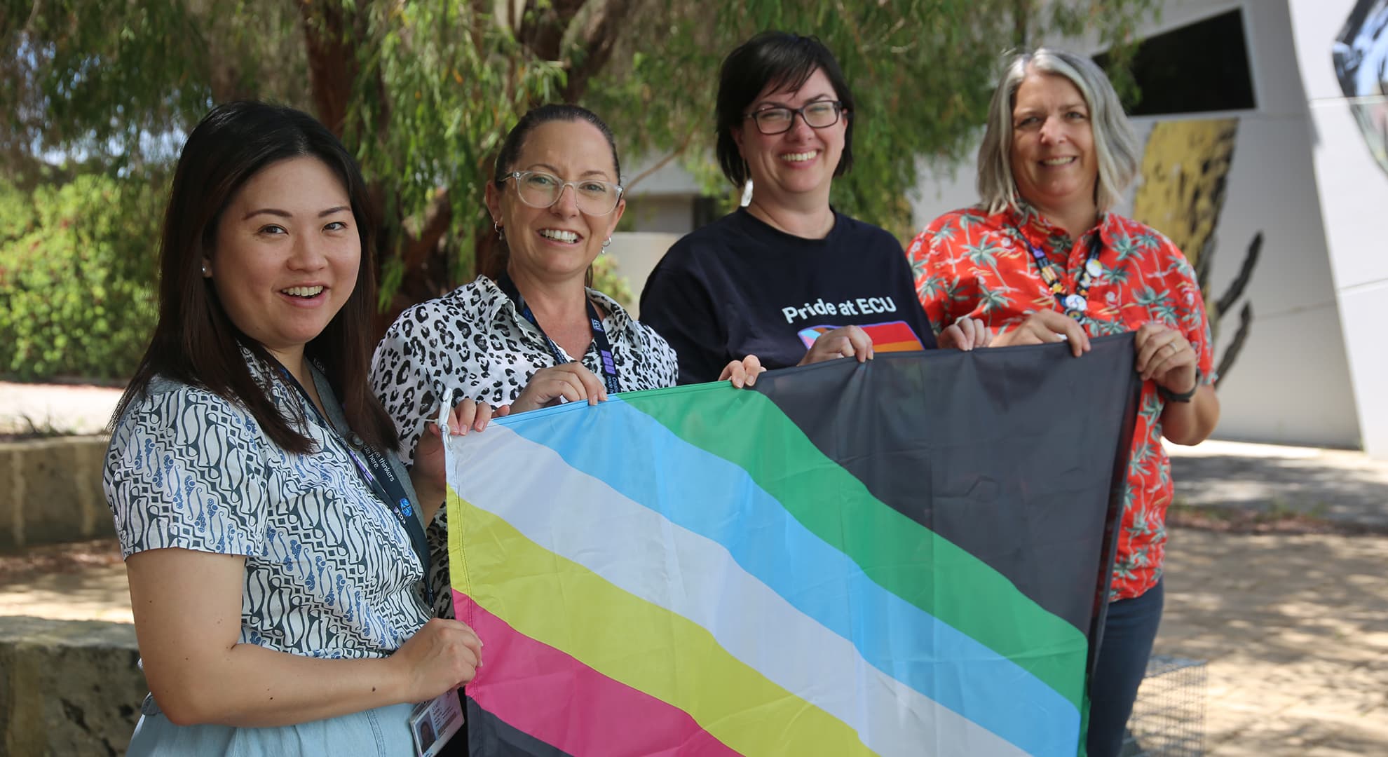 Four ECU staff members hold up the disability pride flag. The flag has grey corners and five diagonal stripes coloured in green, blue, white, yellow and red.