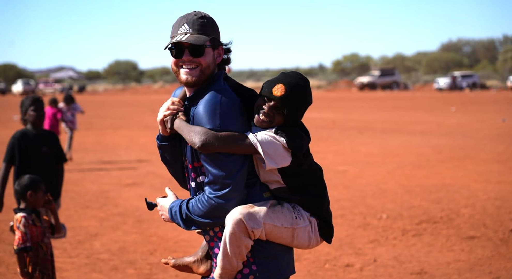 red desert, young guy in baseball cap and sunnies and a beard smiling at camera while young kid in black beanie with orange logo hugs him in a piggy back