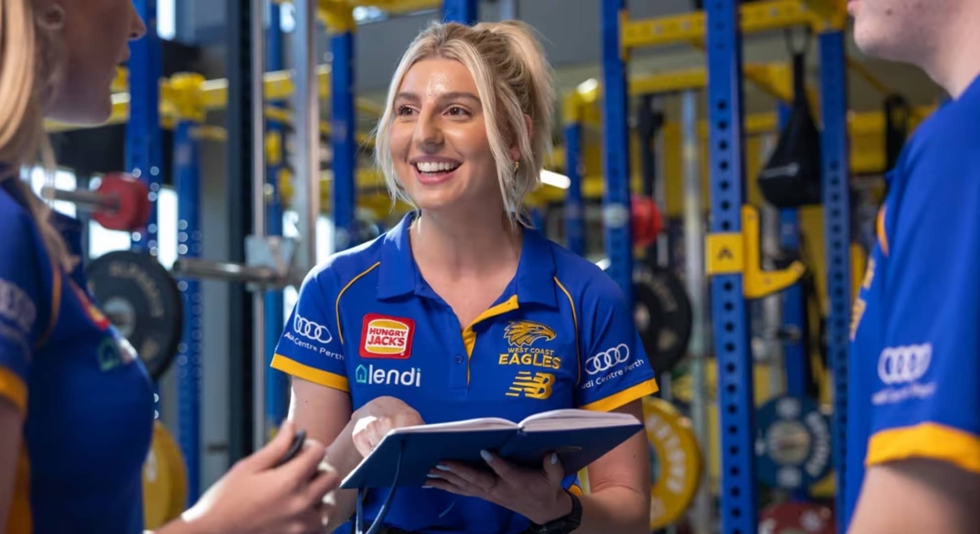 Young blonde woman with hair up in Eagles blue and yellow polo shirt in centre talking to two other people in Eagles blue and yellow polo shirts at edges of photo in a gym with blue and yellow equipment in background