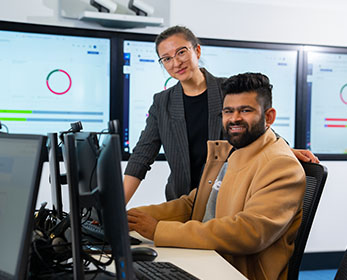 A student and lecturer sit and stand respectively at a computer terminal in a high-tech classroom setting, looking at the camera.