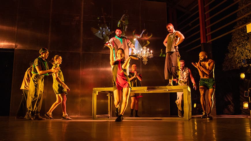 A theatre performance on stage, a woman angles her arms in movement while other people watch on in encouragement.