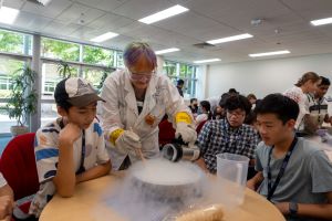 Several teenagers sitting at a table while an adult in a lab coat leans near them to pour liquid nitrogen into a bowl. A cloud of what looks like steam pours from the bowl onto the table while the teenagers look into the bowl.