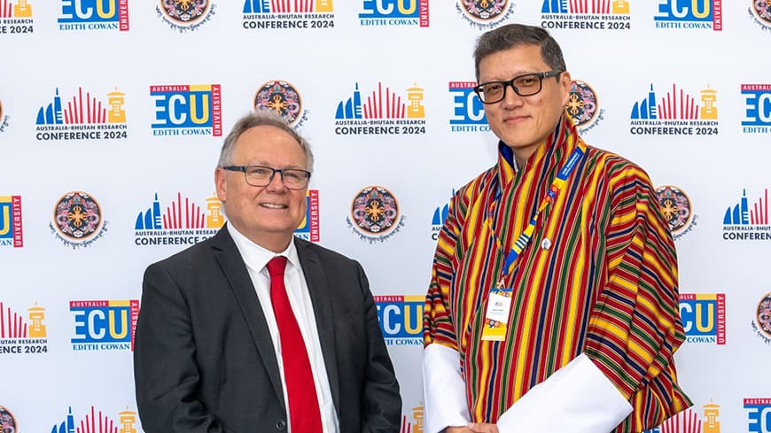 Two people standing in front of media wall with ECU logo, one in a suit and one in traditional Bhutanese dress