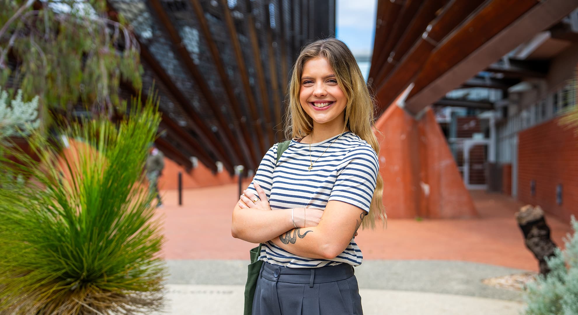 Imogen Haines, an ECU student who's wearing a striped shirt and black pants grins with her arms crossed.