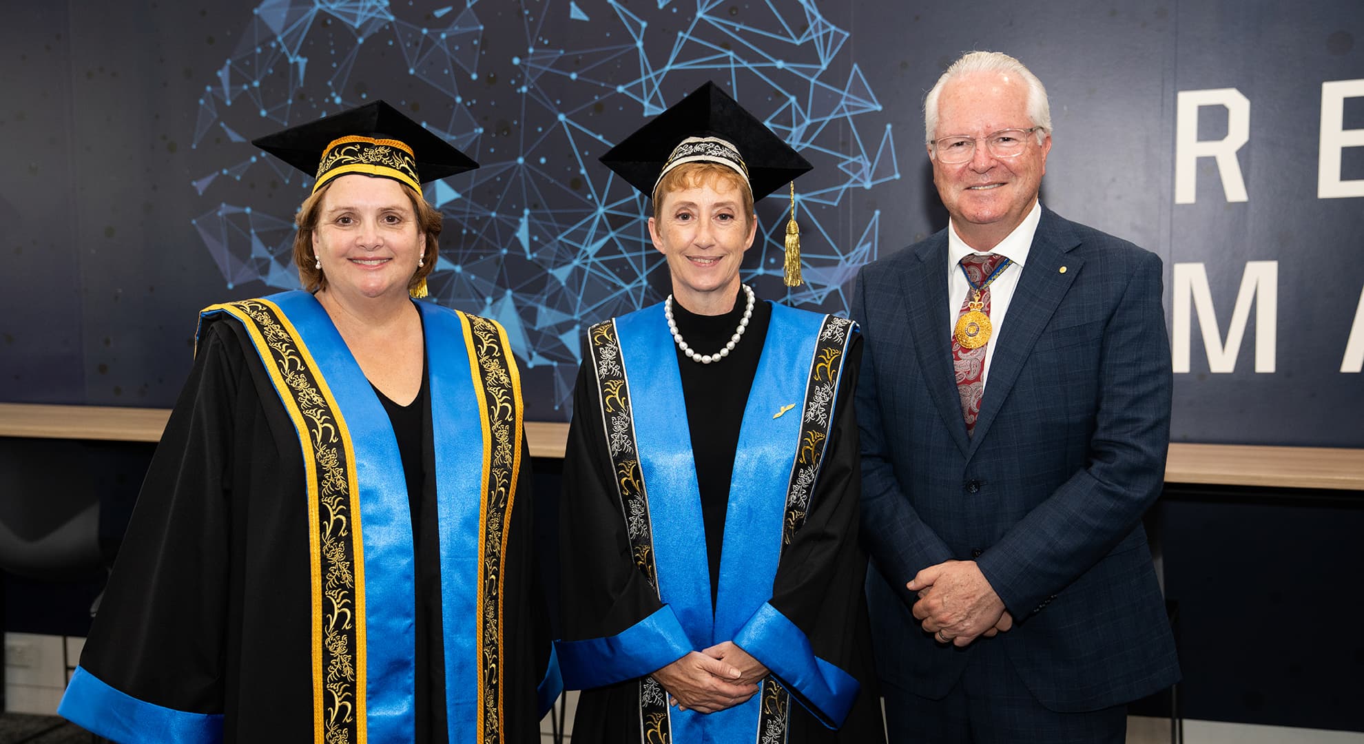 Three people standing in a row, two in university regalia and one in a suit