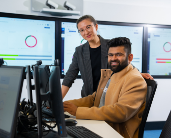 A woman in a grey jacket is standing next to a man in a beige coat working in a computer labe 