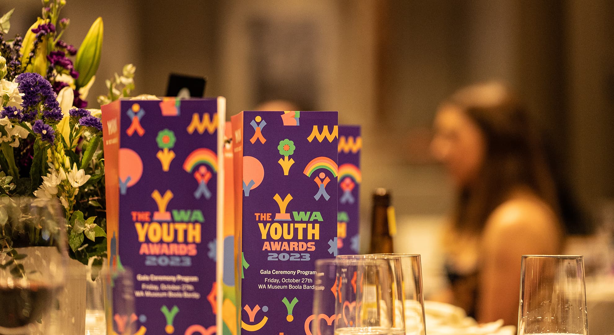 A table setting at the WA Youth Awards 2023 with glasses, flowers and colorful event programs.