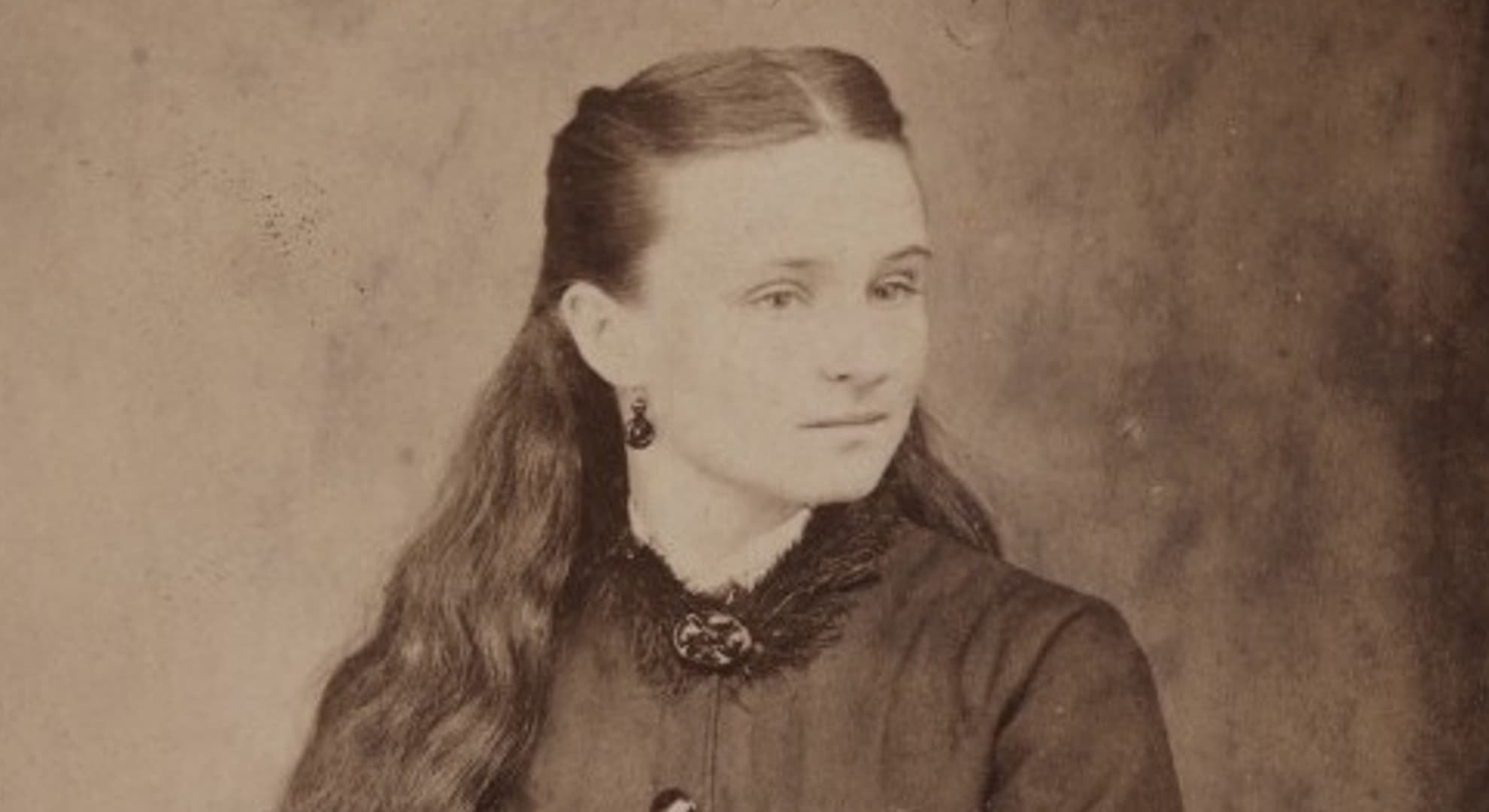 Old sepia toned photograph of head and shoulders of young woman looking to the right, in late 1800s black high-collared dress, with long wavy hair