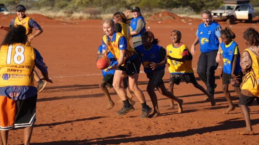 red desert all female, all ages footy game blue and gold uniforms, young woman with blonde braid in centre with the footy