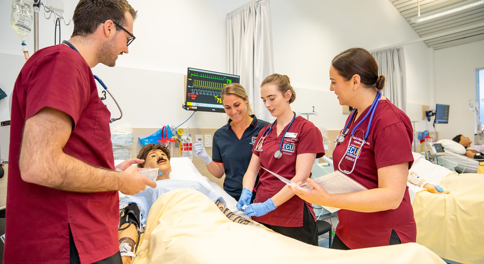 Nursing students and staff standing around a hospital bed with a moustached dummy.