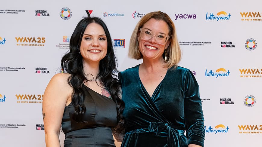 Casey Moller wears a satin black gown, she has curly black hair and tattoos on her arm, she stands next to Hannah Beazley who wears a dark green velvet dress and glasses.