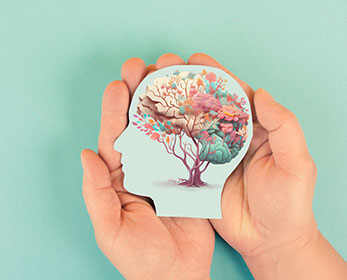 Image of hands holding a paper cut out figure of a human head and brain on a blue background, the brain is depicted as a tree in with leave of blue, white, orange and pink.