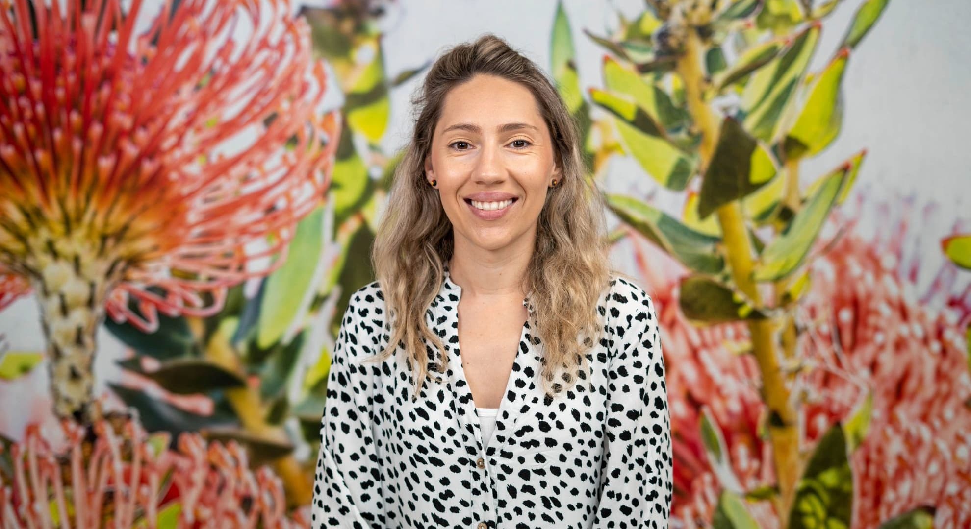 Head and shoulder portrait photograph of smiling woman with light brown wavvy hair past her shoulders, a white blouse with black spots against a backdrop of oversized banksia flowers and leaves