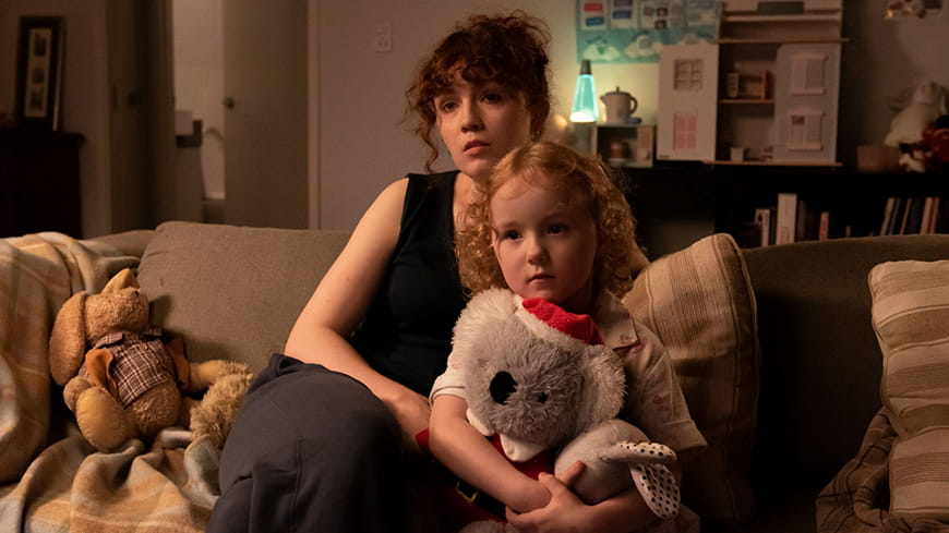 Dark curly haired Mum with little girl with strawberry blonde curly hair sitting cuddled on a couch in a lounge room, little girl is holding a stuffed koala with a Christmas hat on.