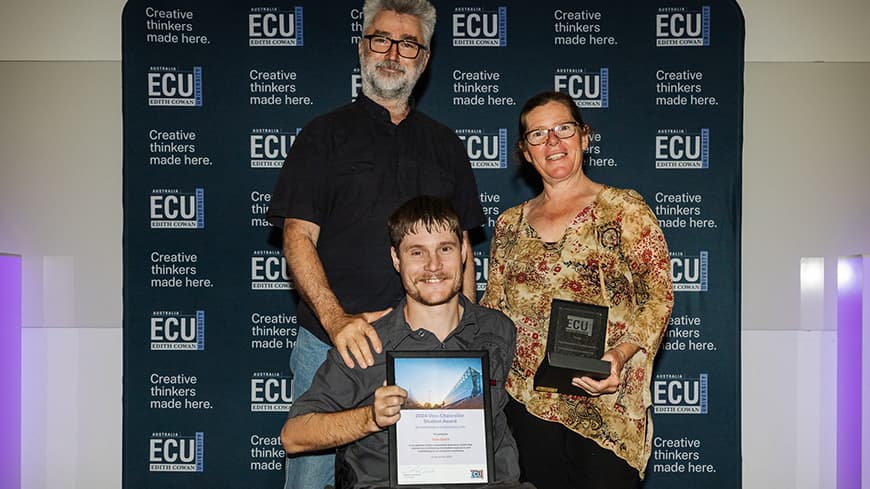 Three people in front of navy blue backdrop with ECU logos, on the left man with silver beard with hand on shoulder of young man in wheelchair in centre, next to woman on right holding an award trophy