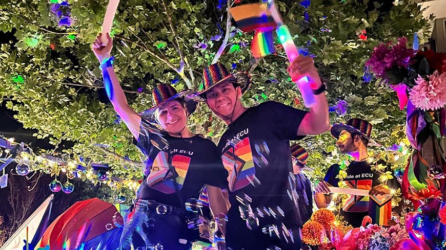 two people arm in arm with matching hats and t-shirts surrounded by colourful flags, bubbles and flowers