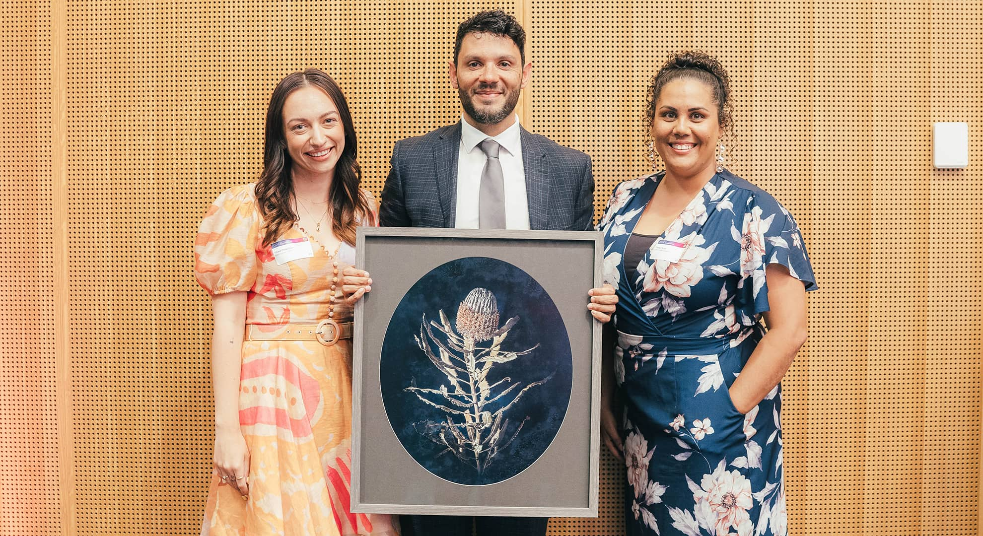 Three people standing in front of a light wood backdrop, man in suit in middle holding a framed artwork of a banksia flower