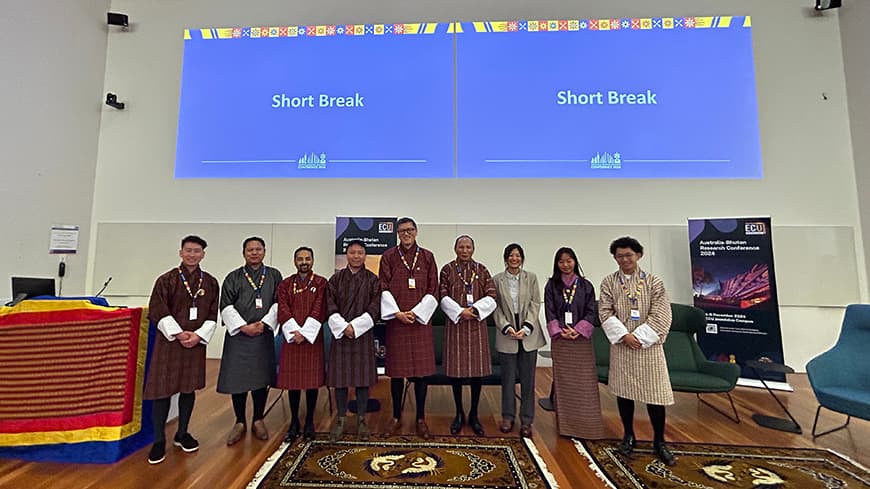 A group of delegates, academics and students dressed in Bhutanese clothing stand at the front of a lecture hall.