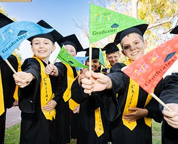 Children's University Primary School Students in their caps and gowns at graduation waving flags and smiling at the camera