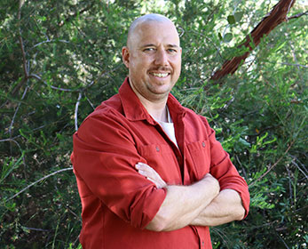 A portrait of Dr David Aldous folding his arms, smiling and standing in from of a tree.