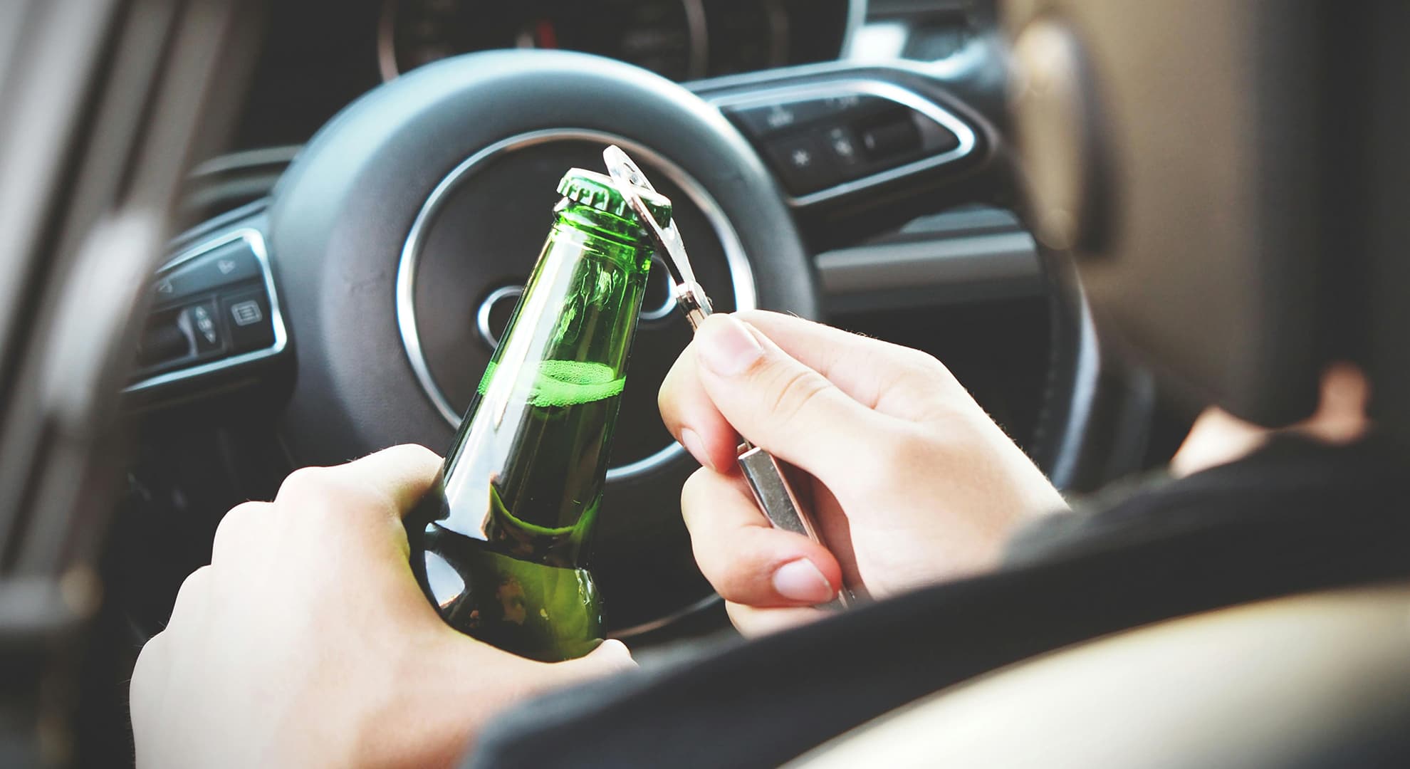 Man behind steering wheel opening a beer bottle.