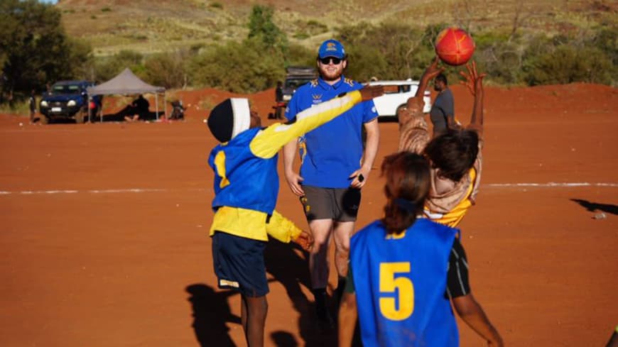 red desert green hills in background, young boys footy game blue and gold uniforms, young man with beard and sunnies and blue cap in centre boys all reaching up for the footy