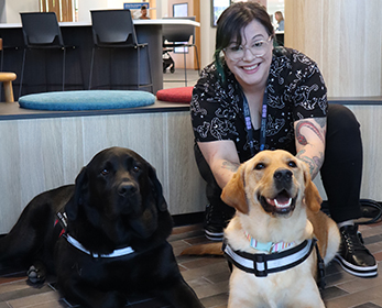 An ECU staff member is smiling at the camera as she pets Edi the dog behind the ears. Watson is also in the frame, and both dogs are looking at the camera.