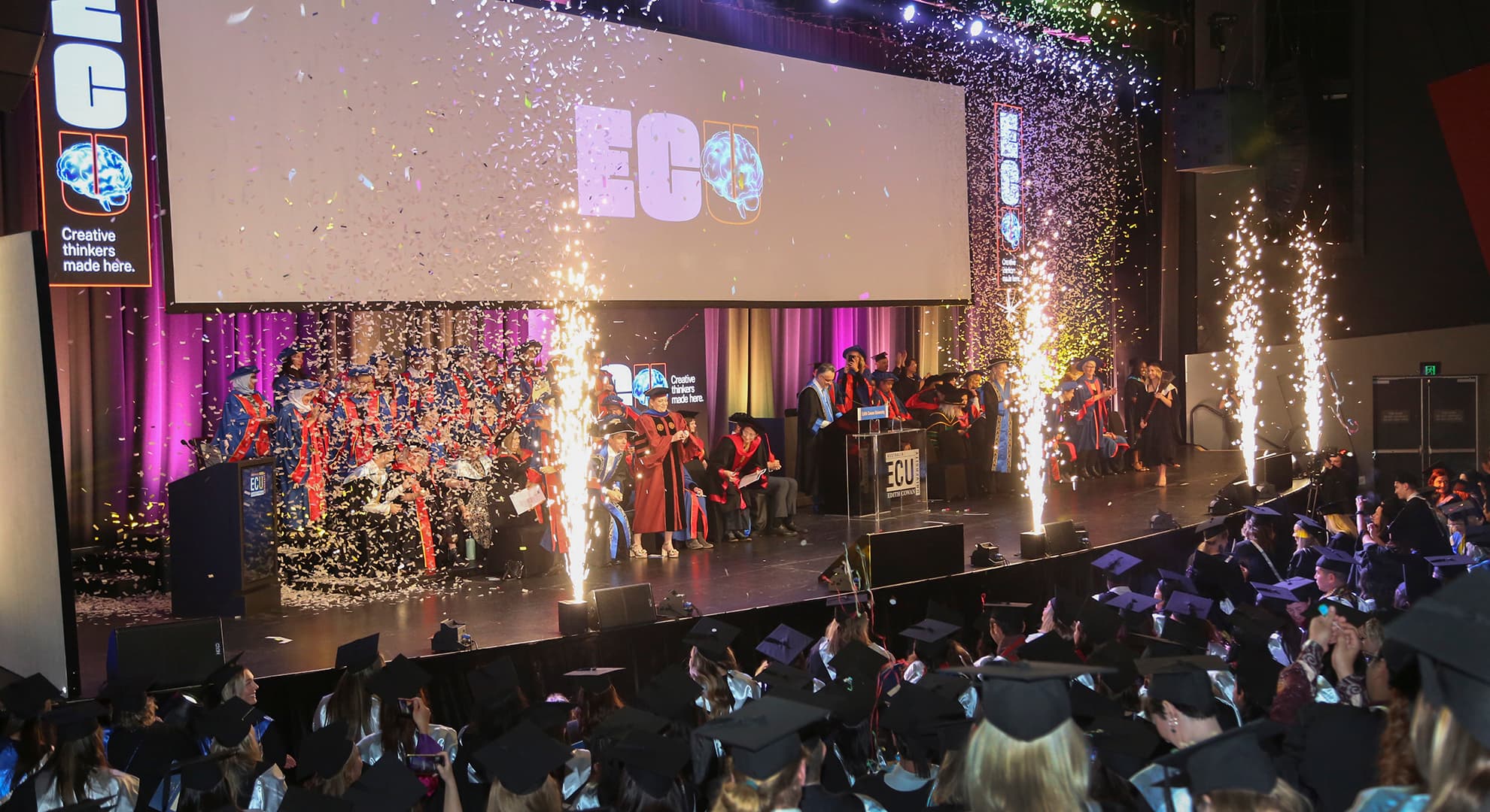 ECU class of 2024 at their graduation ceremony, everyone is dressed in their best with regalias, gowns and caps, there are fireworks up on the stage.