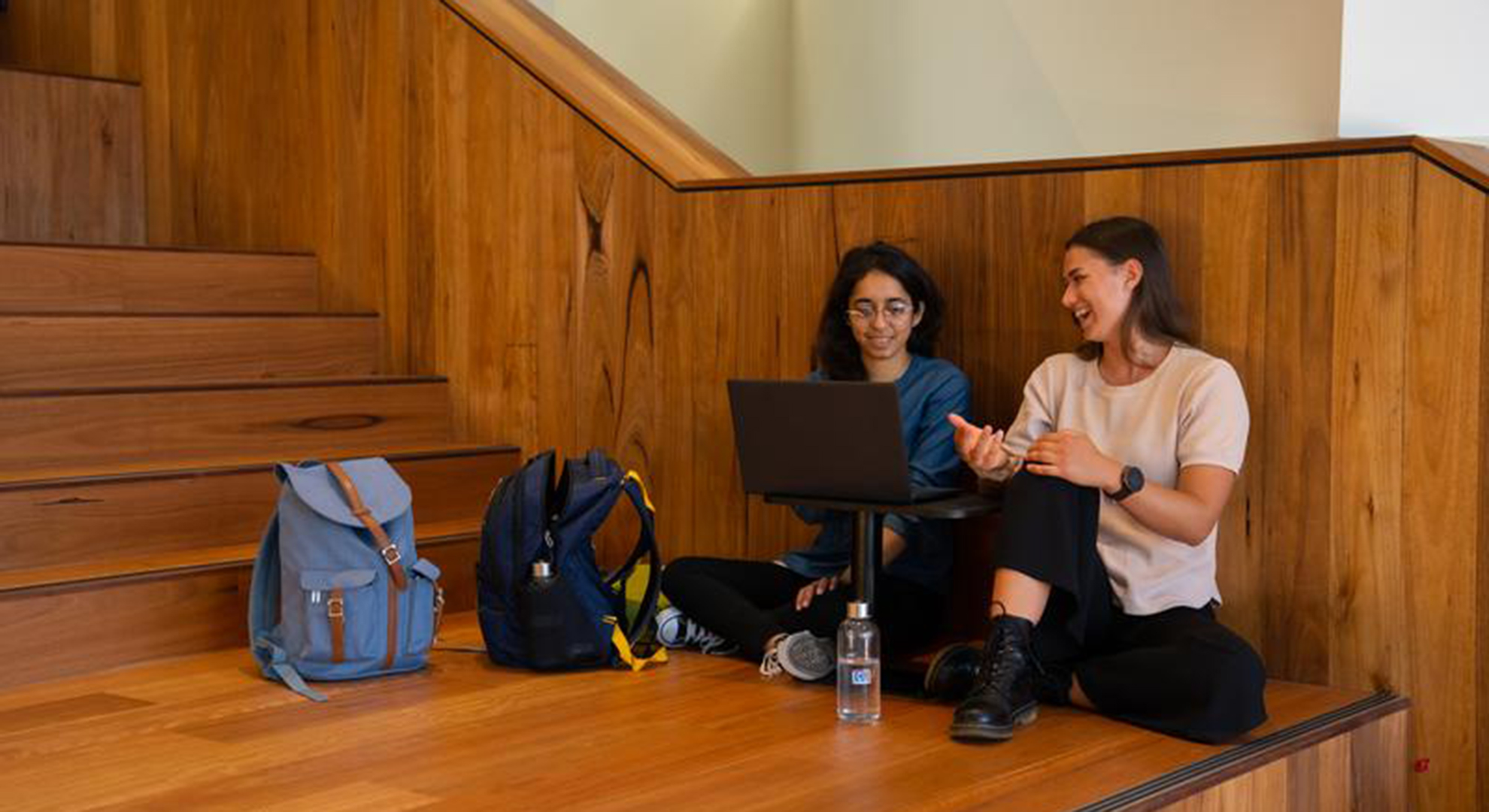 Two students sitting against the wall on the wood panelled Library stairs with back packs