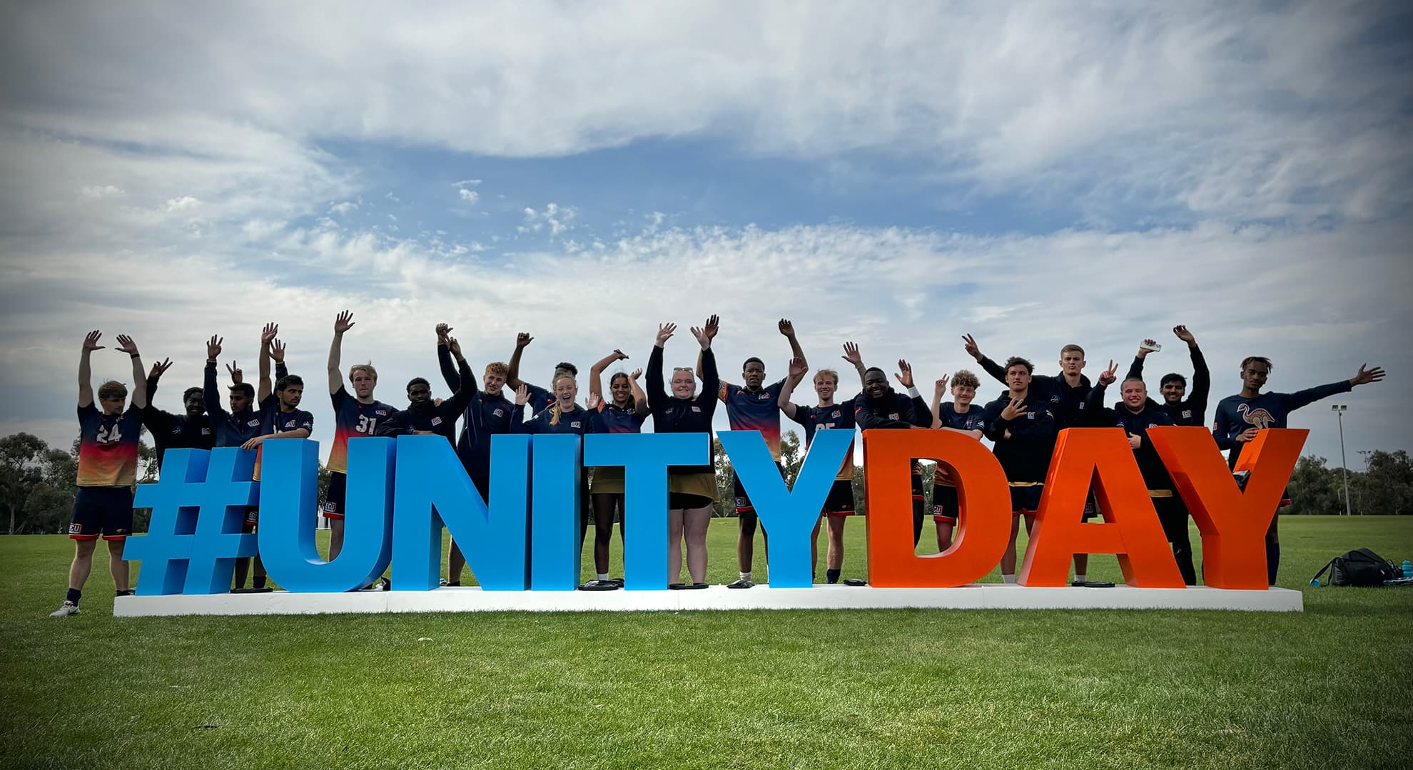 grass oval, giant 3D letters spelling out #UNITY DAY with 20 students in ECU sports uniforms standing behind letters with their arms above their head in celebration