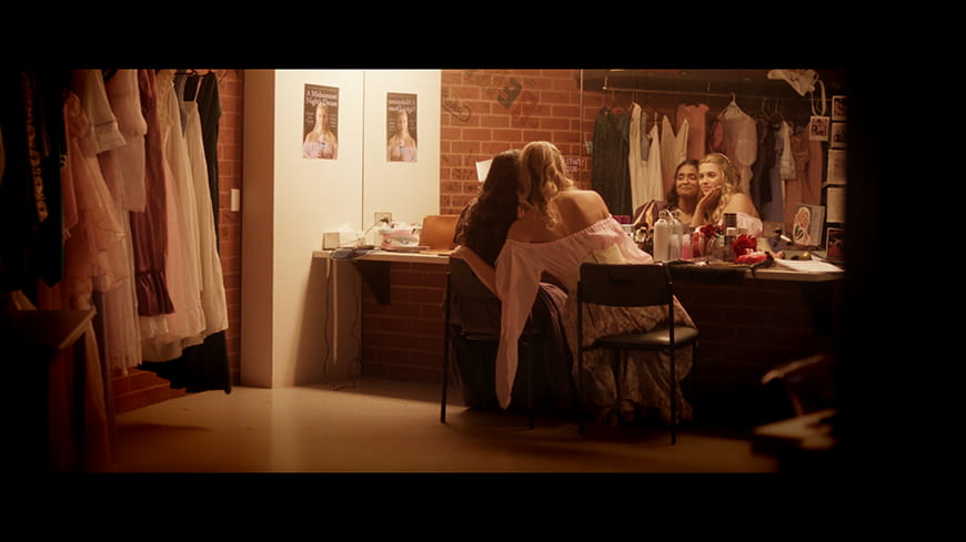 Two actresses cheek to cheek sitting in an backstage dressing room looking in the mirror, one with hand on the other's face.