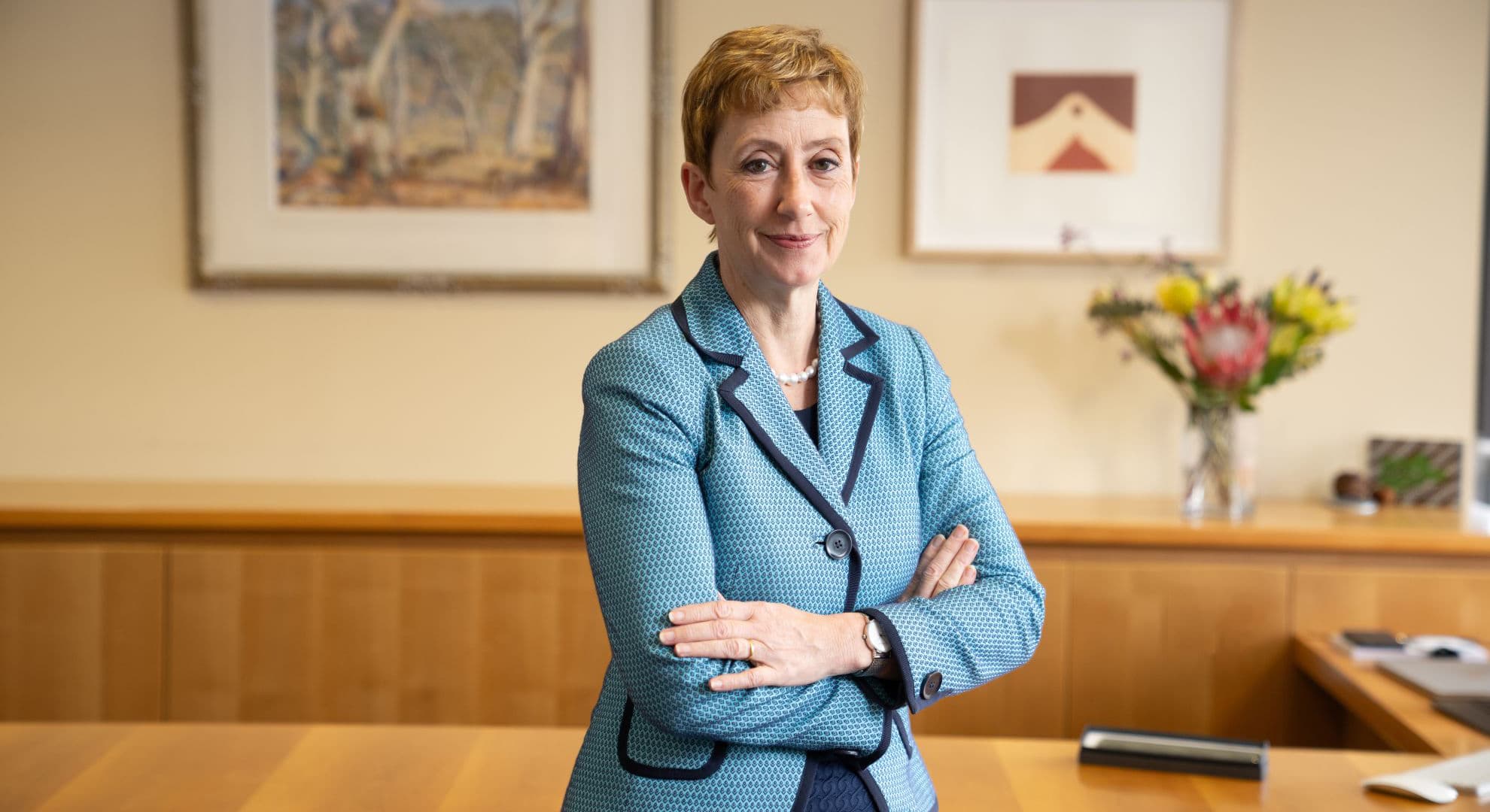 Smiling business woman with short auburn hair and light blue jacket sitting on light wood desk in front of two paintings and native floral arrangement.