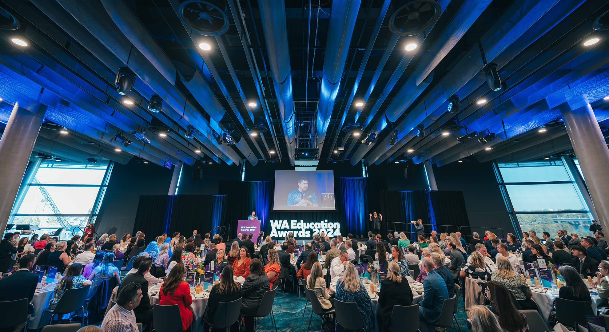 An event space filled with a lot of people sitting around tables in front of a stage.