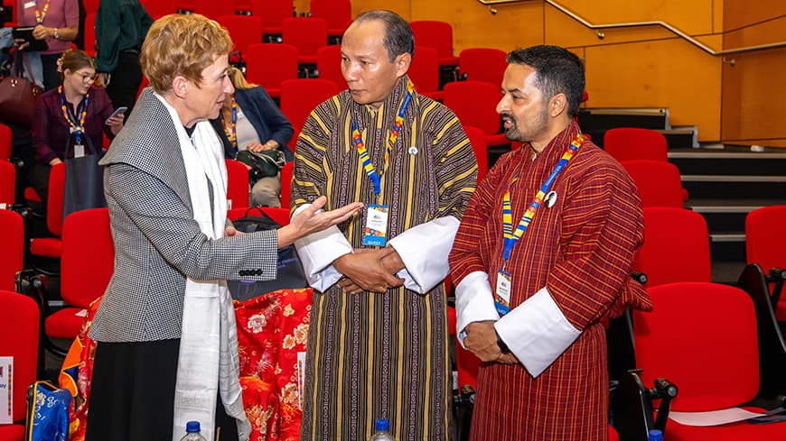 Three people standing in lecture theatre behind a table of covered with colourful cloth and food, two people in traditional Bhutanese dress