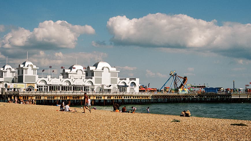 People basking in the sunshine on a shingle beach by South Parade Pier