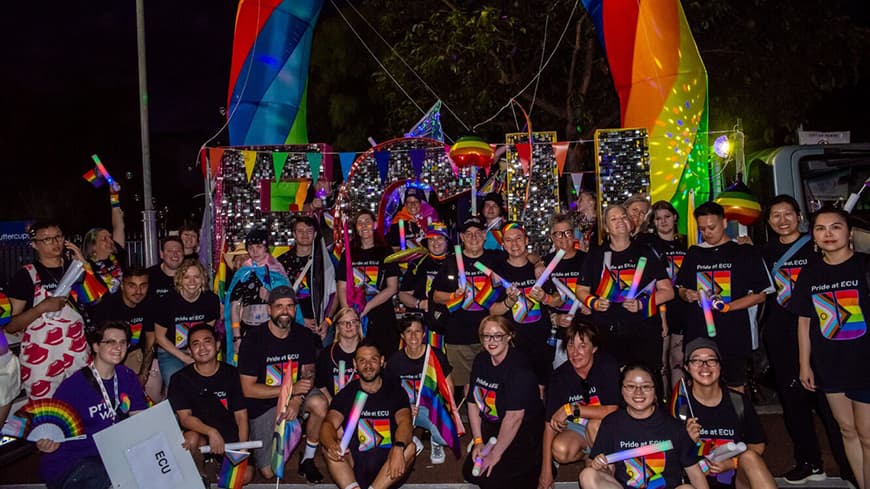 Group photo at night in front of rainbow arch with people in ECU Pride t0shirts waving little rainbow flags