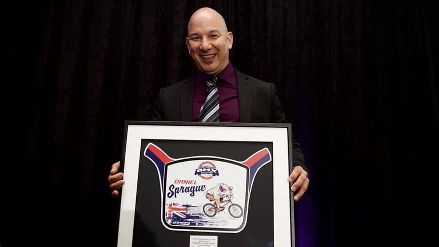 Daniel Sprague wears a suit and tie and holds a framed BMX Hall of Fame award.