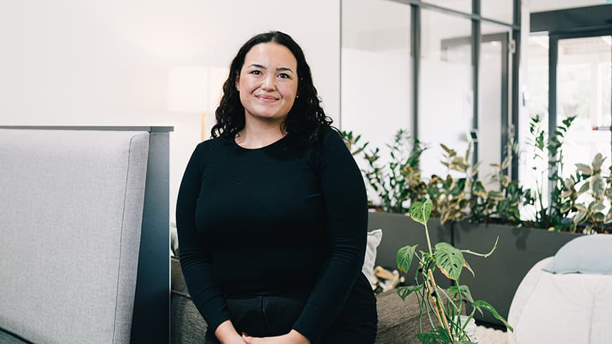 Kathleen Fabre has dark curly hair and wears a black long-sleeved top. She sits in an airy co-working study space.