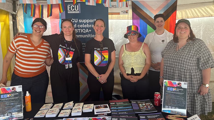 6 people standing behind desk in a stall with rainbow flags