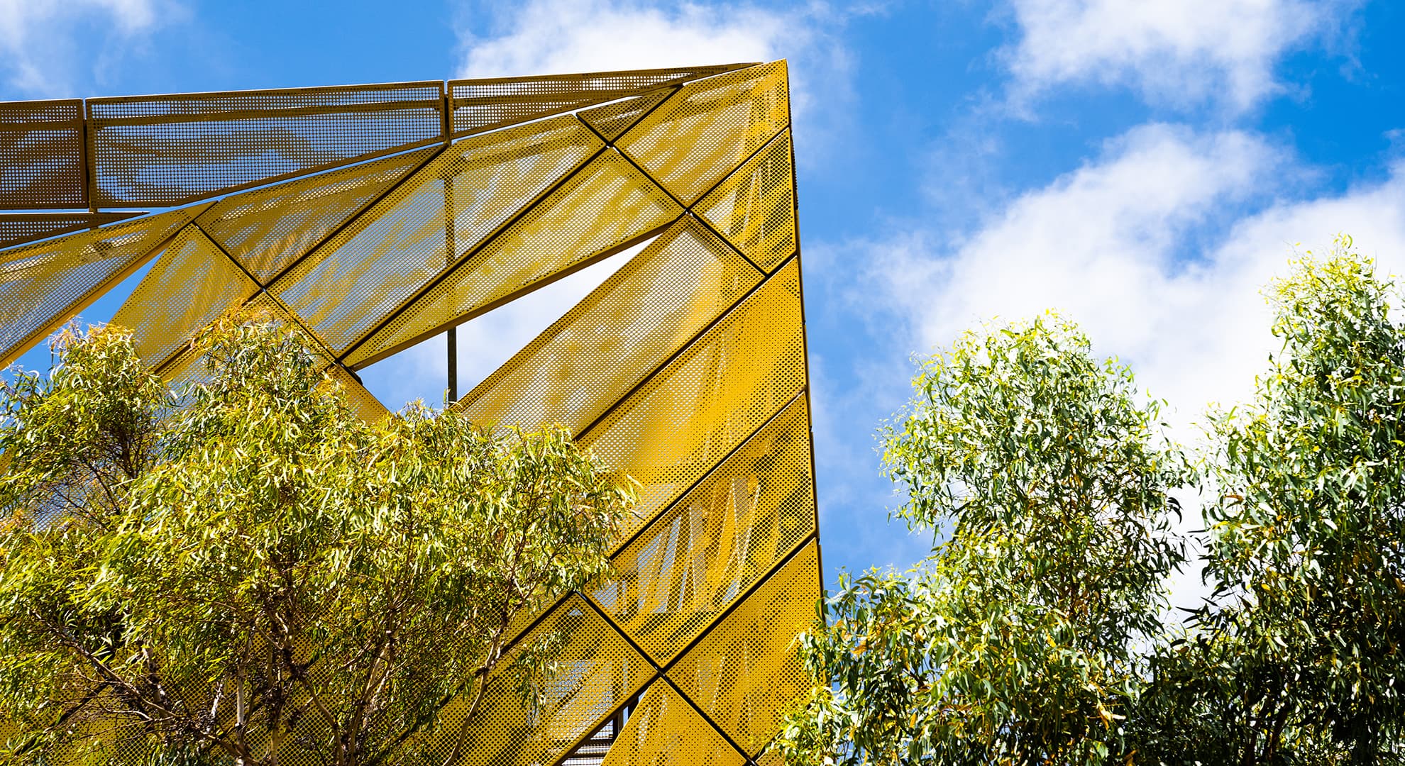 A corner of a gold structural building against the blue sky and clouds and gum trees.