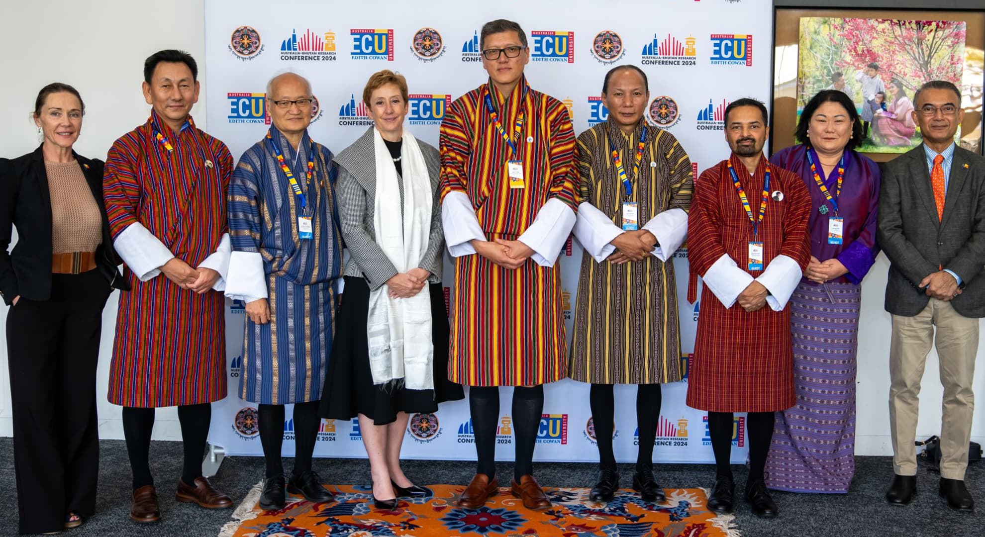 9 people, 5 in traditional Bhutanese dress, standing in a row in front of a media wall with ECU logo