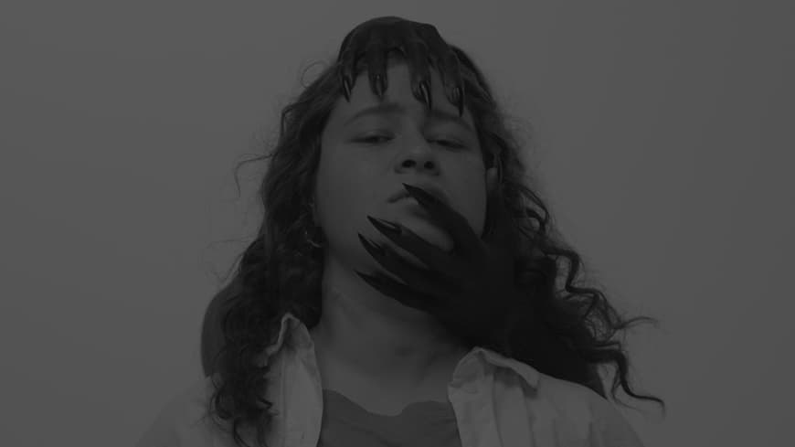 A black and white film still of a young woman, there are demonic hands clutching her face.