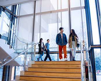 Students walk down the staircase at Mount Lawley Library. Light stream in large glass windows behind them.