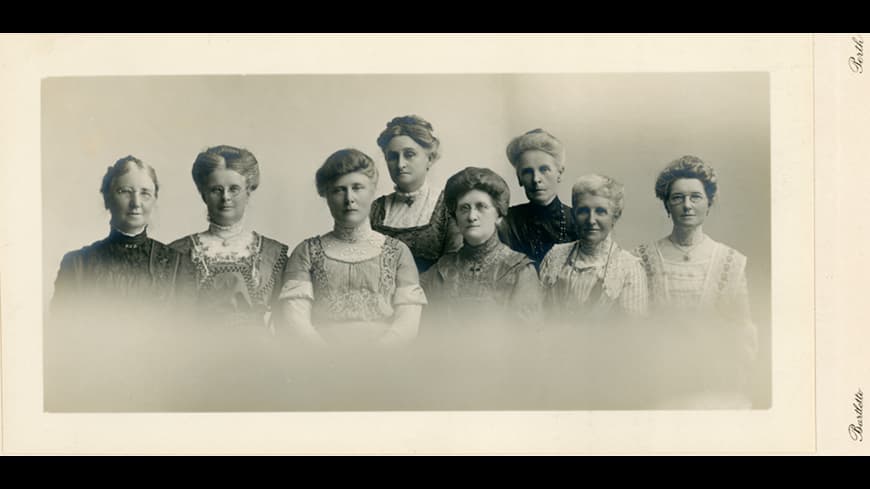 Old sepia toned photograph of a group of 8 women standing in two rows, in ealry 1900s dress, high-collared dresses and hair in soft buns