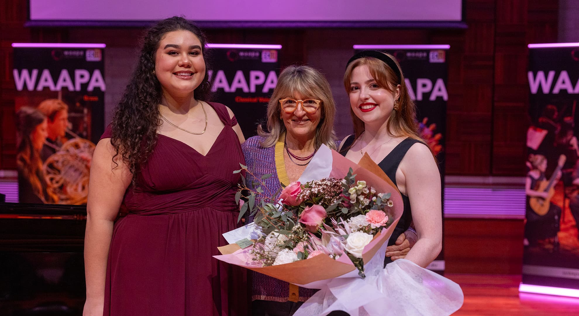 Two WAAPA students dressed in formal wear hold a bouquet of flowers with a director of a philanthropic organisation on stage.