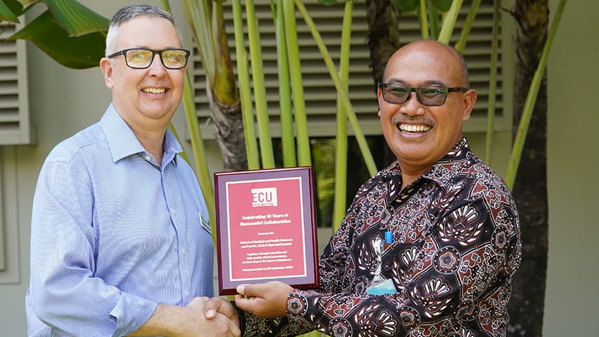 Dr David Reid wears a blue button-down and glasses, he accepts a plaque from Dr. I Gusti Ngurah Ketut Sukadarma, Executive Director of Operational Services at Prof. dr. I.G.N.G Ngoerah Hospital.