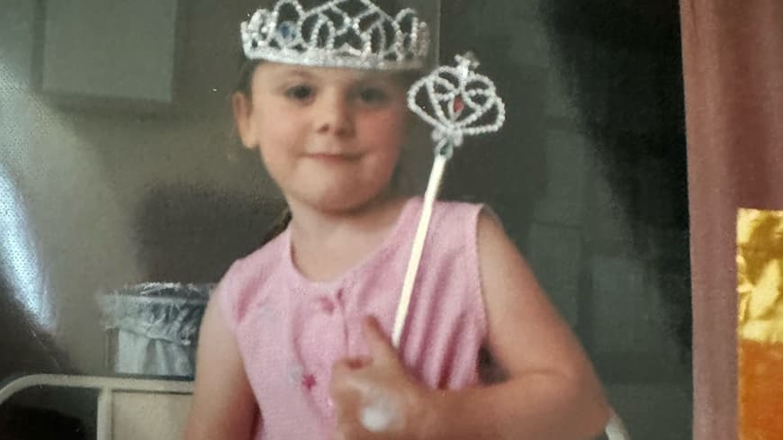 Little girl in pink dress, with plastic silver crown and wand sitting on a  hospital bed with a hospital band on her wrist and a bandaid on her hand