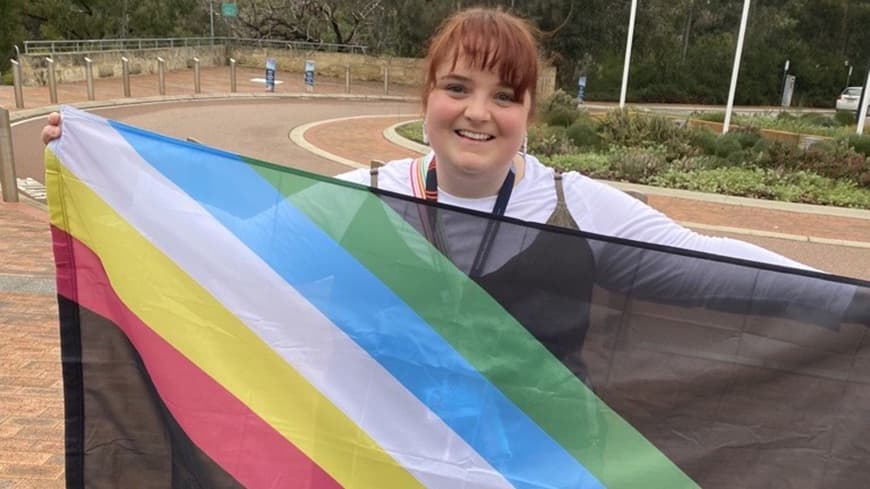 Person standing outside hold the Disability Pride flag in front of them.