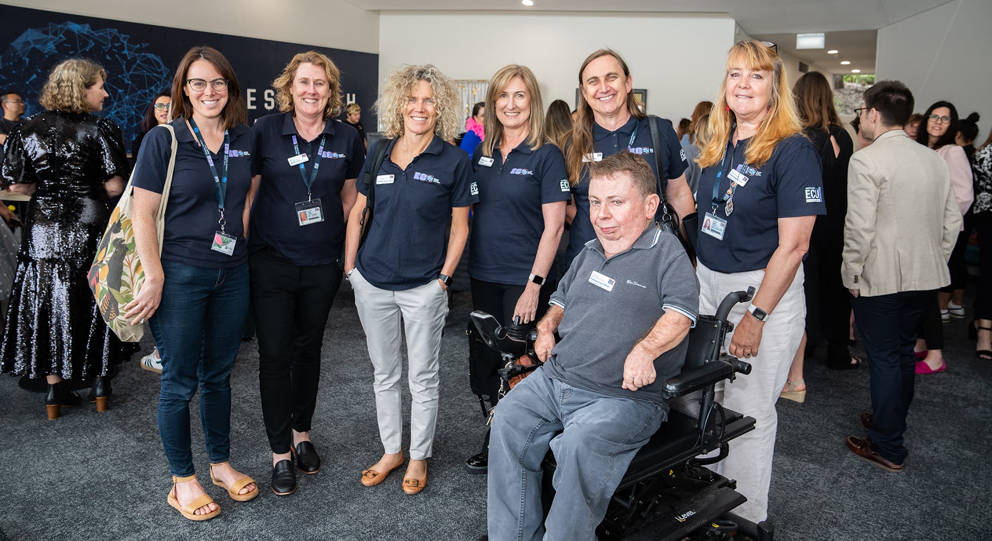 Seven ECU staff members wear navy polos and name badges, they are smiling for the camera, six of them are standing with one staff member in a wheelchair.