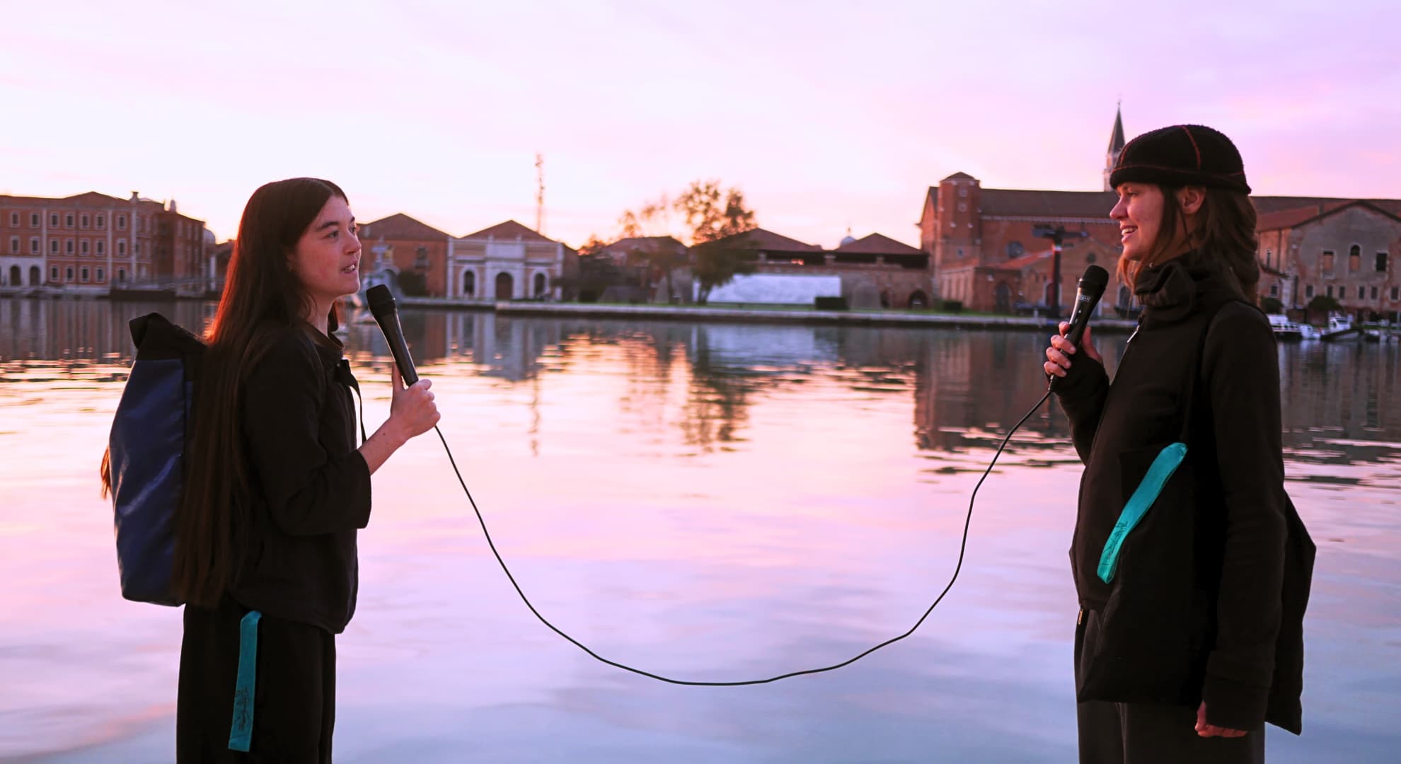 two people in profile holding microphones joined by one cord standing in front of lake surrounded by old European buildings